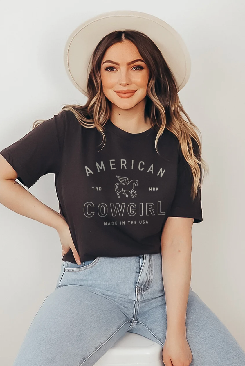 Black t-shirt with the text "American Cowgirl" in a western-style design, perfect for showcasing country pride and rodeo style. Girl wearing cowboy hat and graphic tee.
