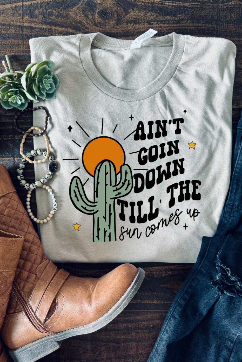 Handmade Bella + Canvas graphic tee with the text "Ain't Going Down Until the Sun Comes Up," perfect for country style and southern charm. Boots and bracelet accessories.