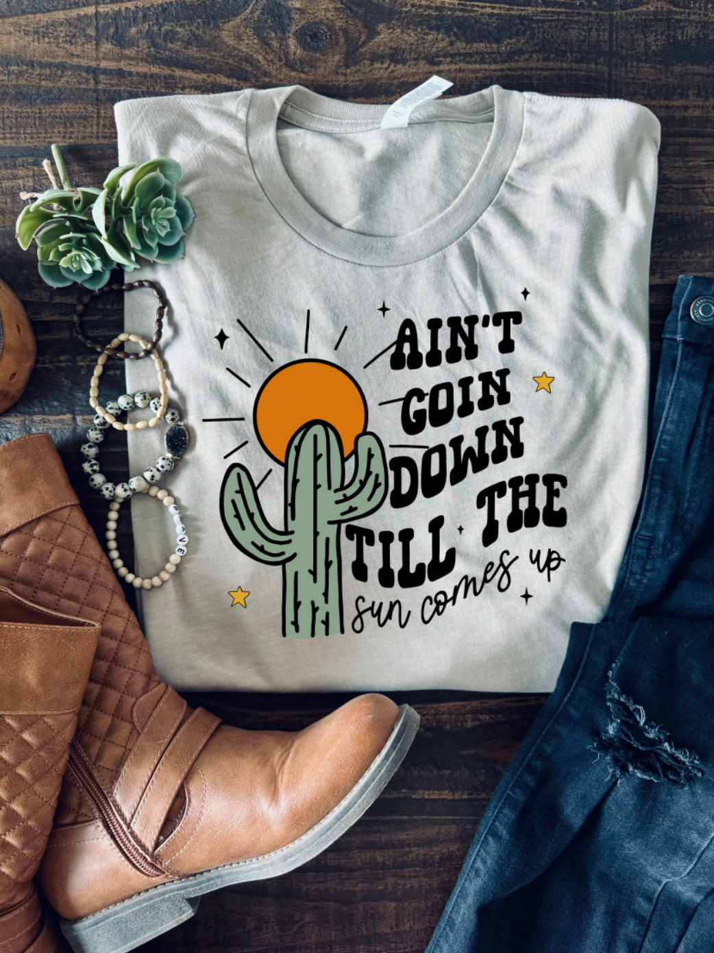 Handmade Bella + Canvas graphic tee with the text "Ain't Going Down Until the Sun Comes Up," perfect for country style and southern charm. Boots and bracelet accessories.
